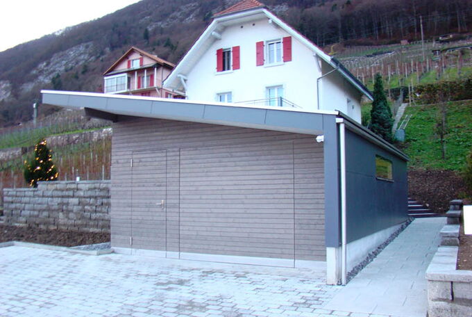 Frontansicht Garage mit Tür und Garagentor in Holzschalung, Seitenwand in Vollkernplatten und Pultdach der Garage Solarzellen eingedeckt